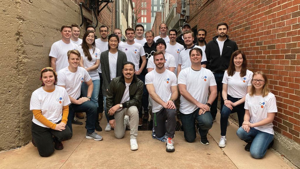 A group of twenty-two people smiling and posing in an alleyway with brick walls. Most are wearing matching white T-shirts featuring a logo. A few are crouching at the front while others stand behind them. Urban buildings are visible in the background.