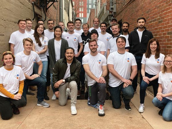 A group of twenty-two people smiling and posing in an alleyway with brick walls. Most are wearing matching white T-shirts featuring a logo. A few are crouching at the front while others stand behind them. Urban buildings are visible in the background.