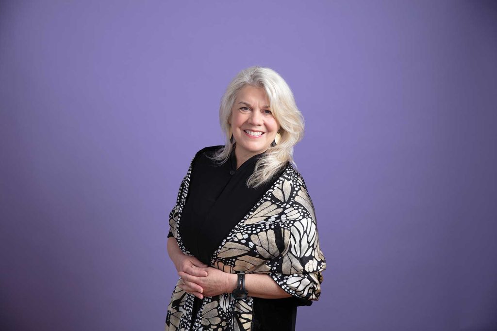 white hair stands smiling against a purple background. Wearing a patterned shawl over a dark outfit