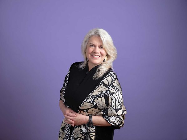 white hair stands smiling against a purple background. Wearing a patterned shawl over a dark outfit