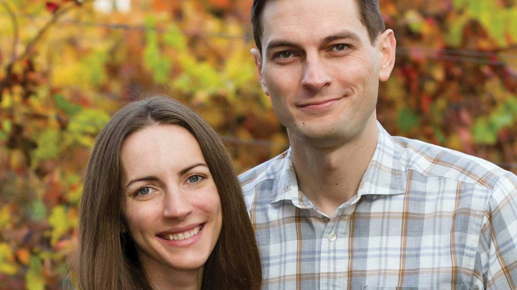 standing outdoors with a background of colorful autumn foliage. The man is wearing a plaid shirt