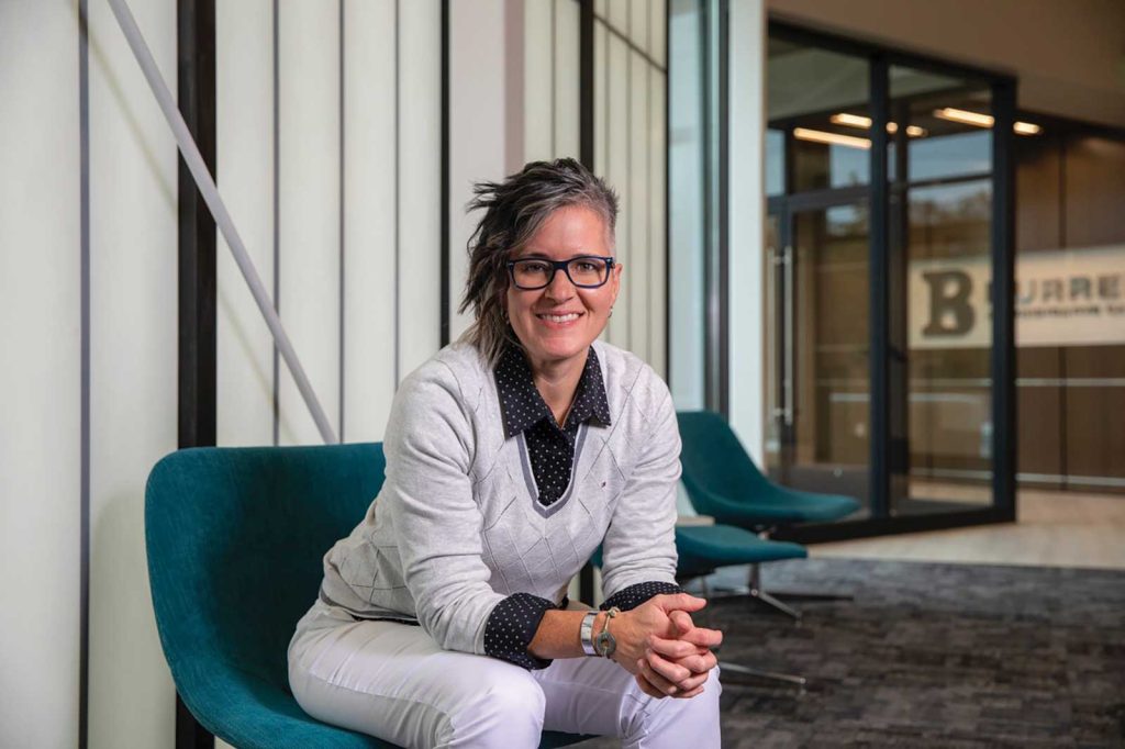 styled hair and glasses sits smiling on a teal chair. They wear a light gray sweater over a black polka dot shirt with white pants. The background features large windows and a glimpse of an entrance.