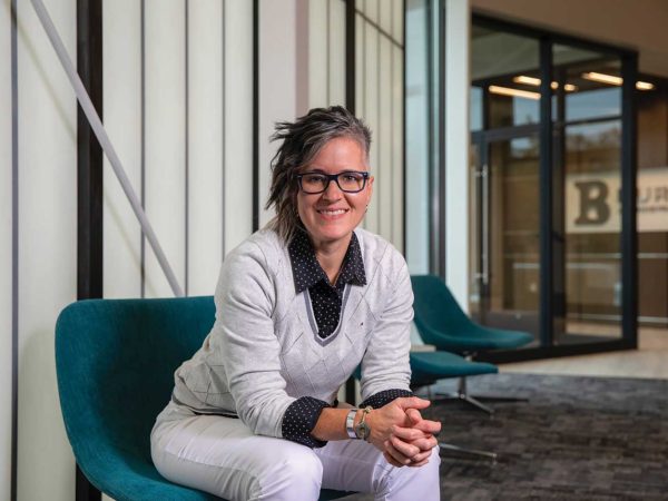 styled hair and glasses sits smiling on a teal chair. They wear a light gray sweater over a black polka dot shirt with white pants. The background features large windows and a glimpse of an entrance.