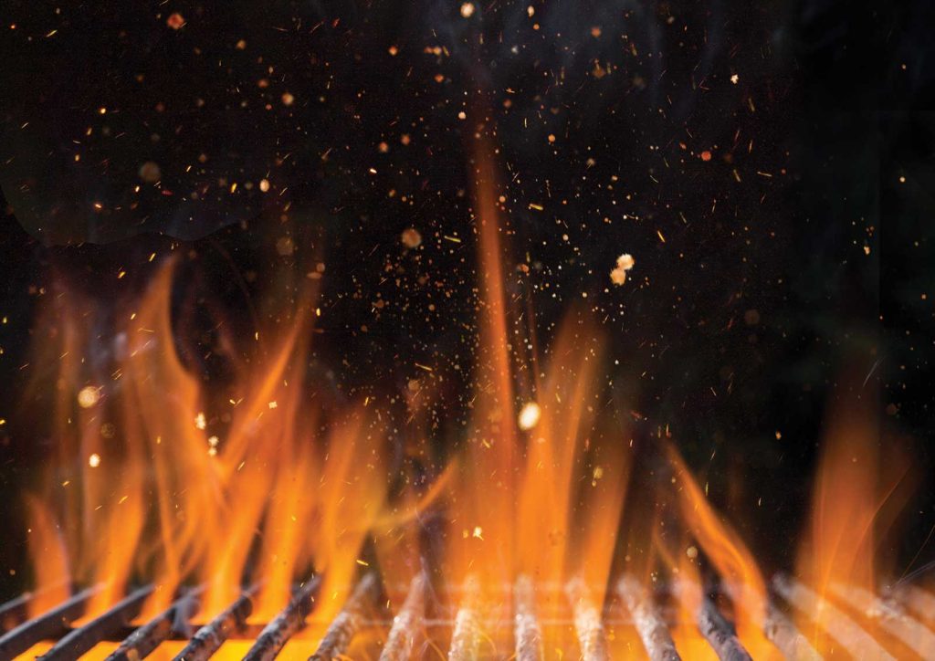 Close-up of an active barbecue grill with bright orange flames and sparks flying against a dark background. The metal grill grates are visible at the bottom.
