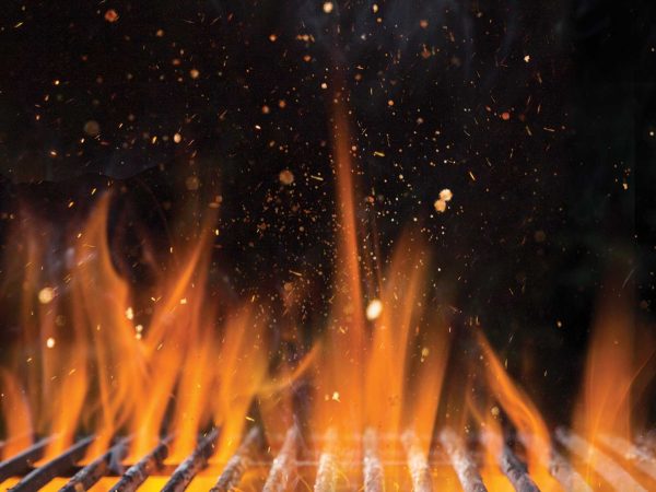 Close-up of an active barbecue grill with bright orange flames and sparks flying against a dark background. The metal grill grates are visible at the bottom.