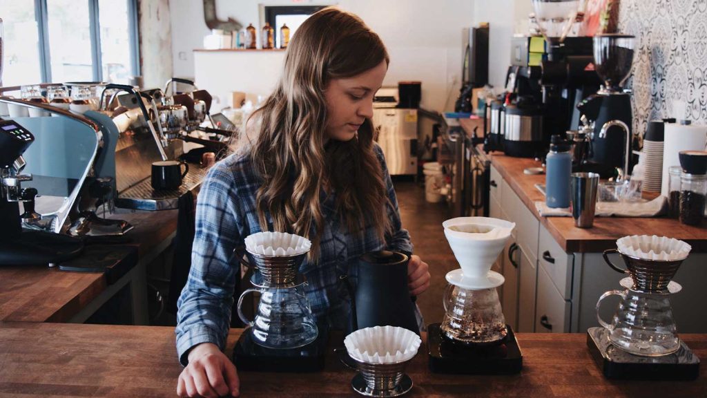 A woman with long hair is preparing coffee in a cozy café. She is using a pour-over coffee setup with multiple drippers on a wooden counter. The background features coffee machines and various café items.