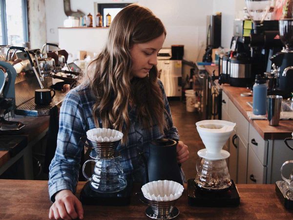 A woman with long hair is preparing coffee in a cozy café. She is using a pour-over coffee setup with multiple drippers on a wooden counter. The background features coffee machines and various café items.