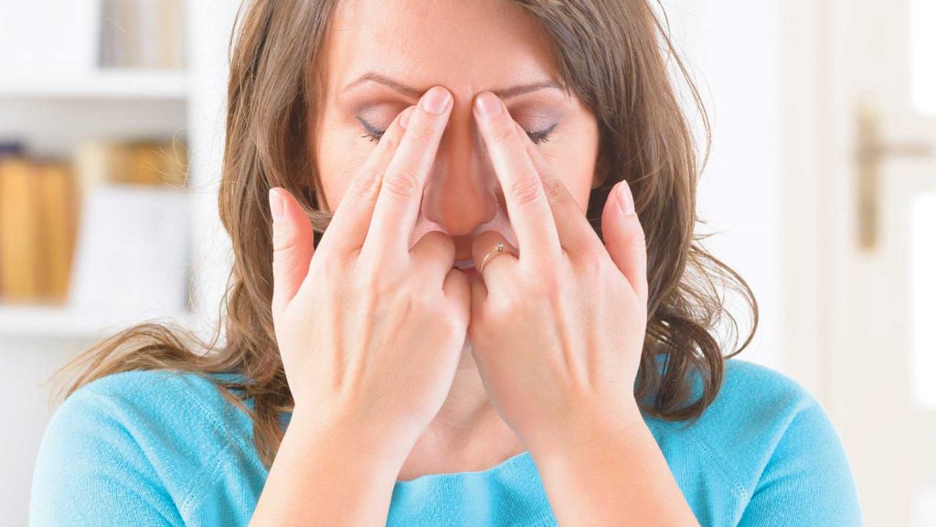 A person with long brown hair wearing a blue shirt is pressing their fingers against their forehead between their closed eyes. They appear to be focusing or relieving tension. Bookshelves and a door are in the blurred background.