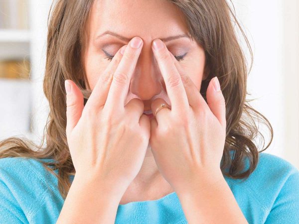 A person with long brown hair wearing a blue shirt is pressing their fingers against their forehead between their closed eyes. They appear to be focusing or relieving tension. Bookshelves and a door are in the blurred background.
