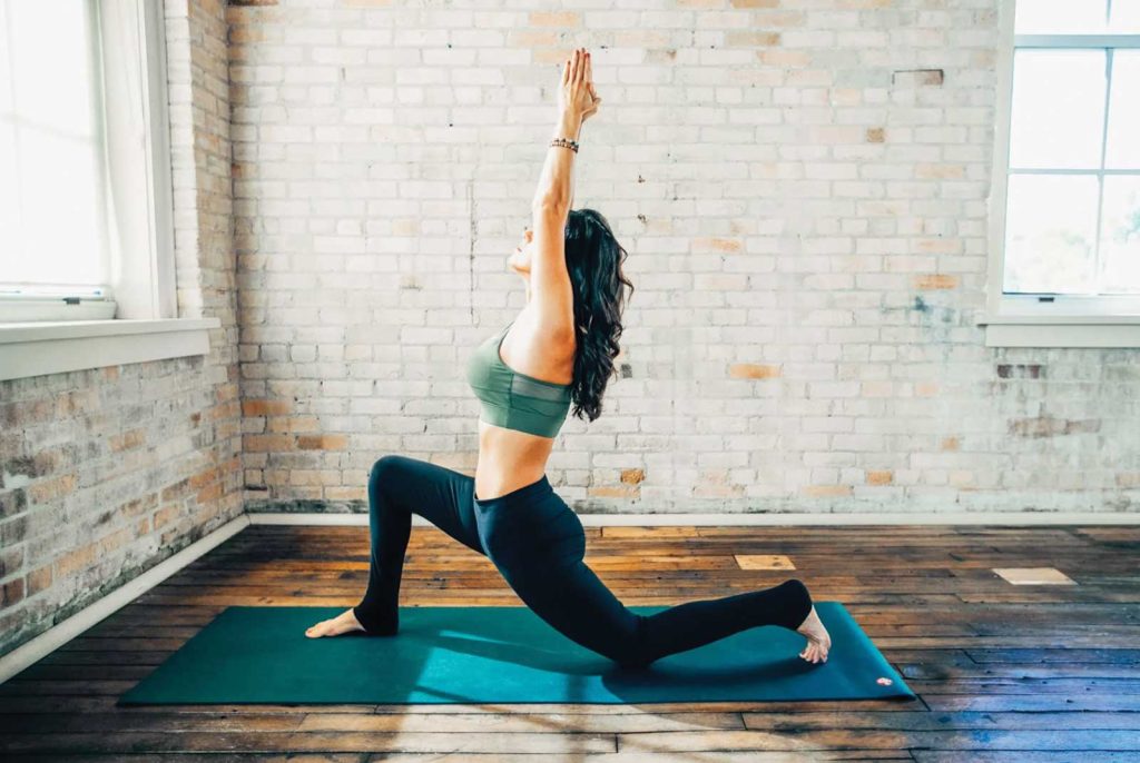 A women in a yoga move on a yoga mat inside of a yoga studio with several large windows and white washed brick walls