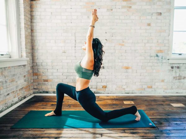 A women in a yoga move on a yoga mat inside of a yoga studio with several large windows and white washed brick walls