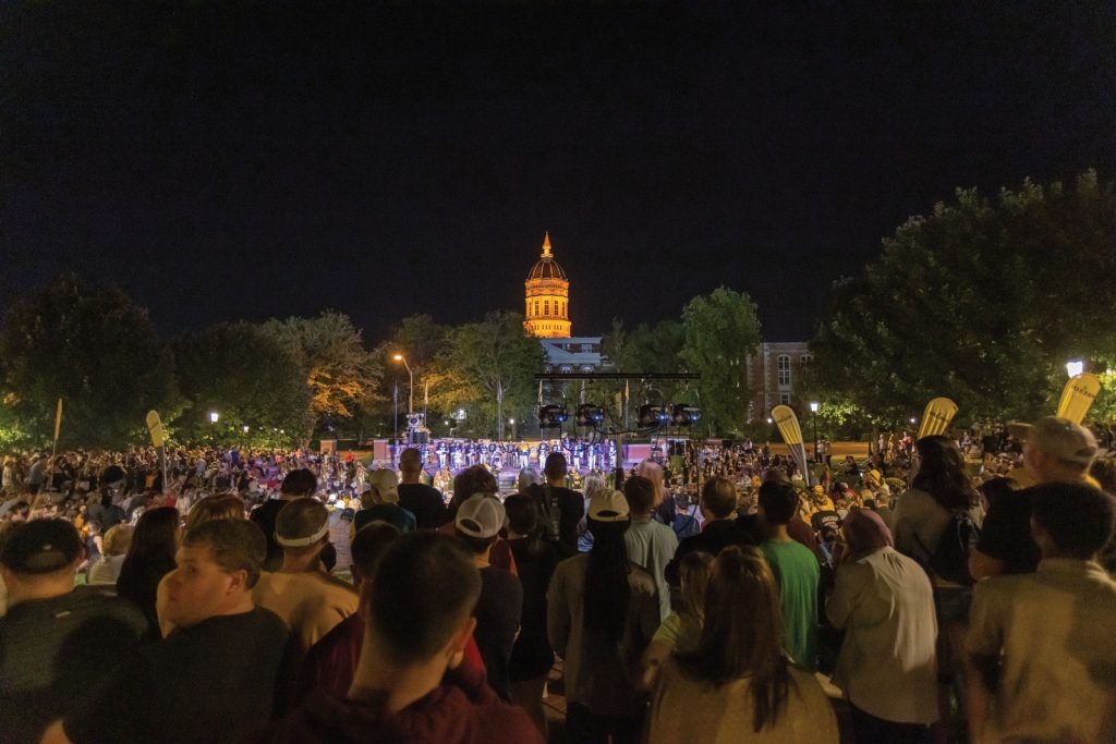 with people facing a lit stage. Trees and additional lighting create a vibrant scene.|People gather outdoors in front of a decorated building with a Homecoming 2021 banner. The scene is illuminated with colorful lights against a dusky sky. Attendees sit on hay bales
