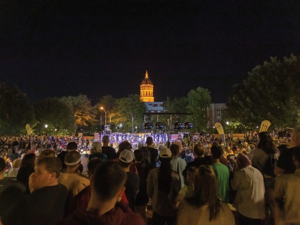 with people facing a lit stage. Trees and additional lighting create a vibrant scene.|People gather outdoors in front of a decorated building with a Homecoming 2021 banner. The scene is illuminated with colorful lights against a dusky sky. Attendees sit on hay bales