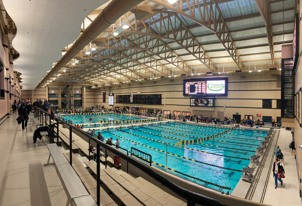 under a high ceiling with skylights. Spectators sit on bleachers