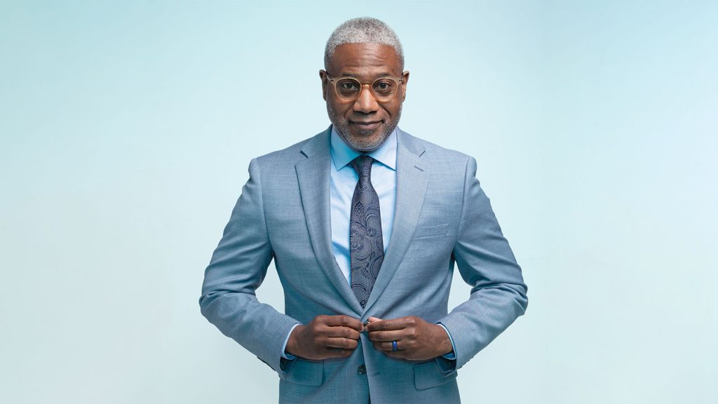 Jr. buttoning the front of his blue suit jacket standing in front of a blue background|A headshot of a man named Dr. Lester Woods