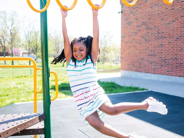 her legs extended. The background features a brick building and trees on a sunny day.|A young child wearing a white shirt and plaid shorts stands confidently on top of a jungle gym at a playground
