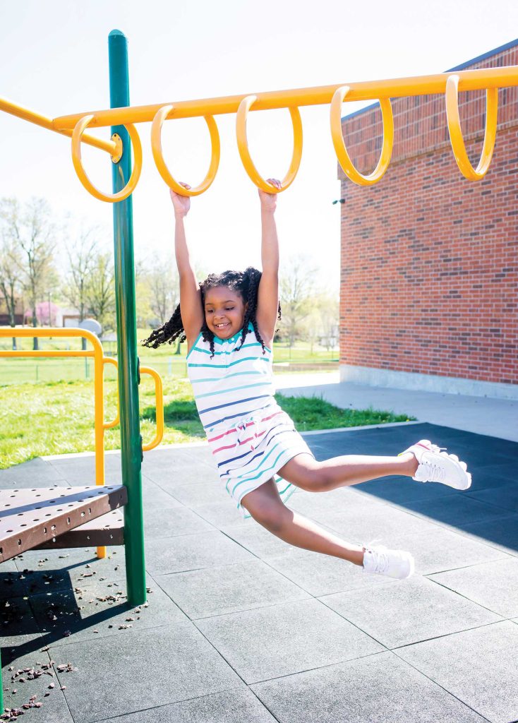 her legs extended. The background features a brick building and trees on a sunny day.|A young child wearing a white shirt and plaid shorts stands confidently on top of a jungle gym at a playground