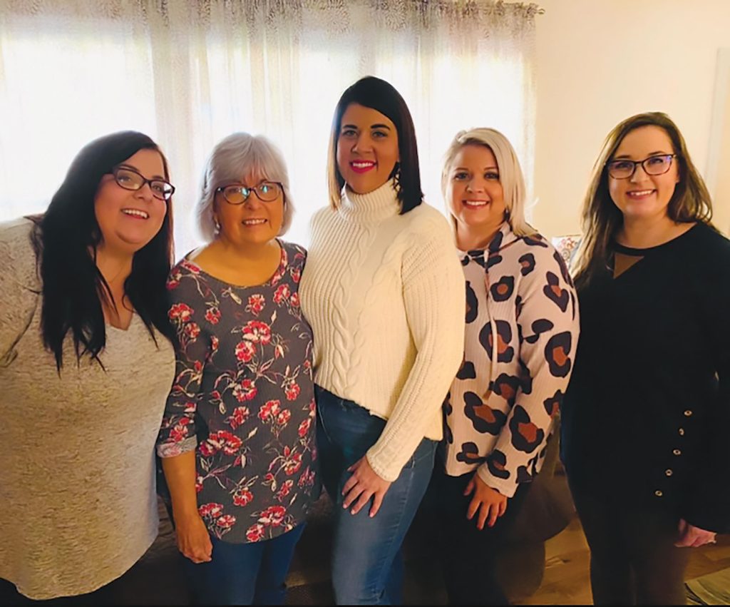 surrounded by younger family members. The backdrop features white shelves with books and decorative items. Everyone is dressed in formal or semi-formal attire