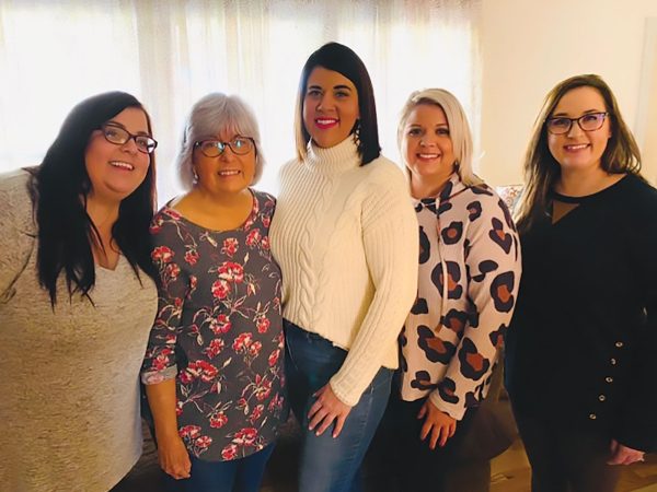 surrounded by younger family members. The backdrop features white shelves with books and decorative items. Everyone is dressed in formal or semi-formal attire