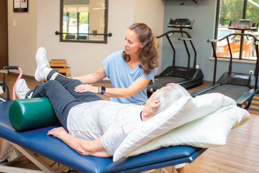 examining the ankle of someone lying down. Exercise equipment is visible in the background of the gym.|A person lies on a therapy bed in a gym