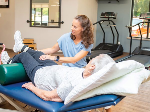 examining the ankle of someone lying down. Exercise equipment is visible in the background of the gym.|A person lies on a therapy bed in a gym