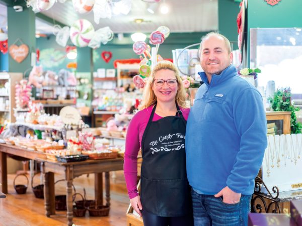 Amy and Mike Atkinson stand inside their shop|A baker lays fresh Carmel into a tray of chocolate in a kitchen at the Candy Factory|Valentines day themed box of assorted chocolate produced by the candy factory|behind the scenes of the candy factory where a tray of caramels is being coated with a layer of chocolate by a automated machine|Owners of the Candy Factory