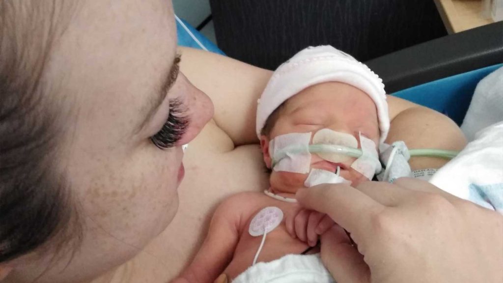 the infant in a pink hat connected to medical tubes. As she gently holds the babys tiny hand