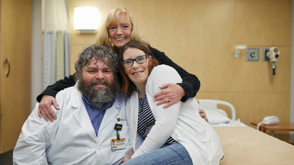 hugging both. The room has beige walls and medical equipment is visible.|A woman wearing glasses and a white outfit stands next to a bearded man in a white medical coat