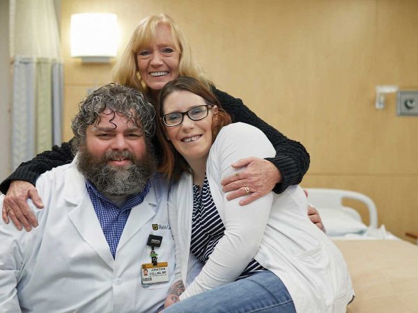 hugging both. The room has beige walls and medical equipment is visible.|A woman wearing glasses and a white outfit stands next to a bearded man in a white medical coat