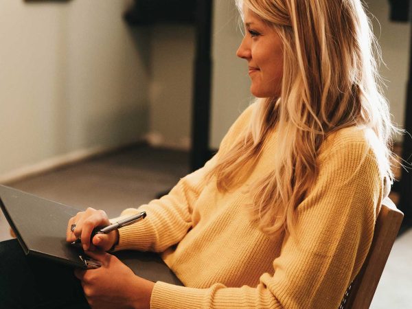 wearing a yellow sweater. She holds a clipboard and pen