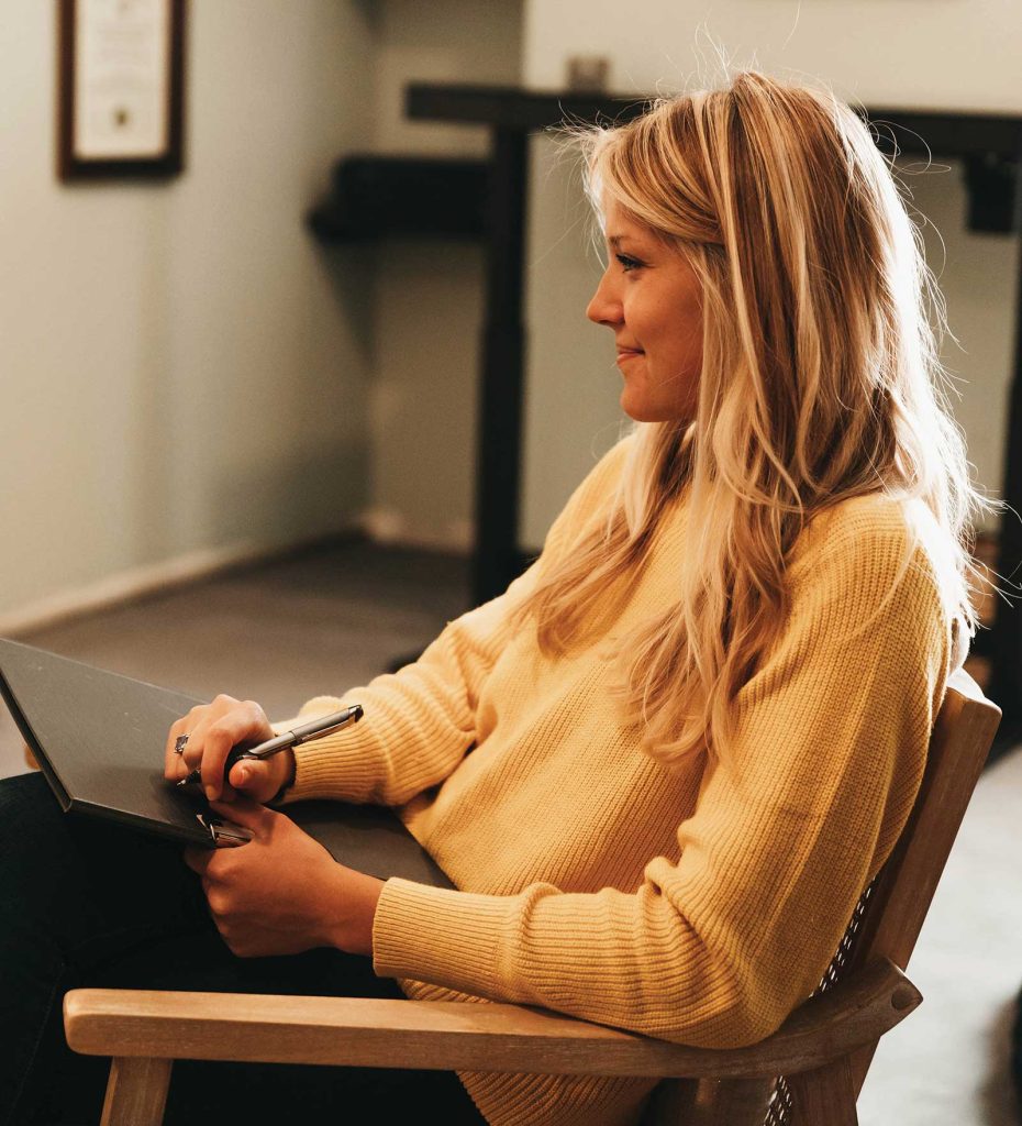 wearing a yellow sweater. She holds a clipboard and pen