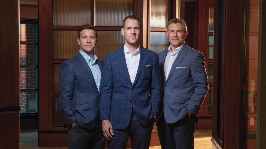 and two have their hands in their pockets. The background features glass-paneled doors and warm wooden tones.|A smiling man in a blue blazer and a checkered shirt stands in front of wooden panel doors. The background features a mix of wood and frosted glass.|A man in a blue suit and white shirt stands in front of a wooden and glass paneled background