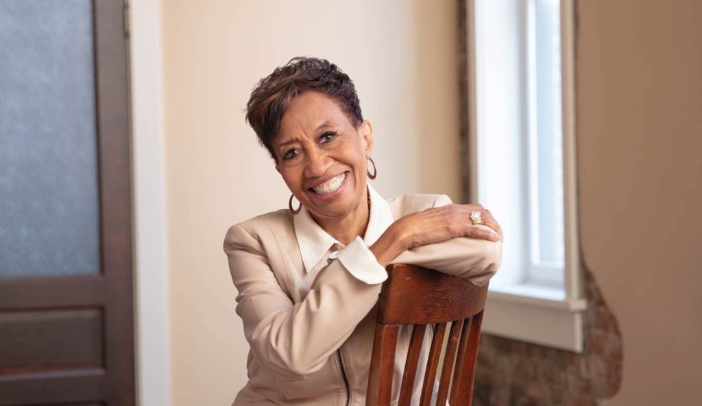 leaning forward with arms crossed over the backrest. She wears a light blouse and jacket