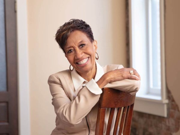 leaning forward with arms crossed over the backrest. She wears a light blouse and jacket
