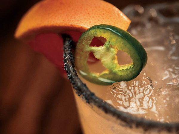 garnished with a jalapeño slice and an orange wedge. The glass rim is coated with black salt. The background is dimly lit