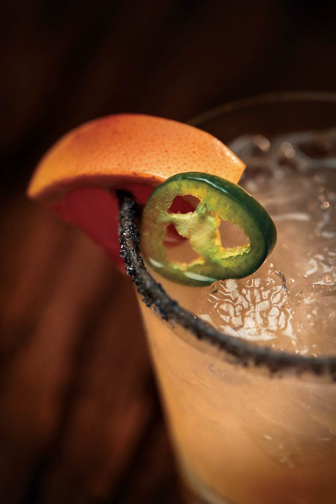 garnished with a jalapeño slice and an orange wedge. The glass rim is coated with black salt. The background is dimly lit