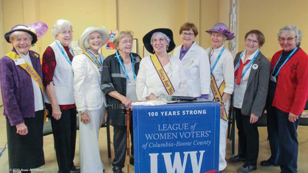 smiling. They are wearing various hats and sashes with the word Votes. They are posed behind a banner that reads 100 Years Strong