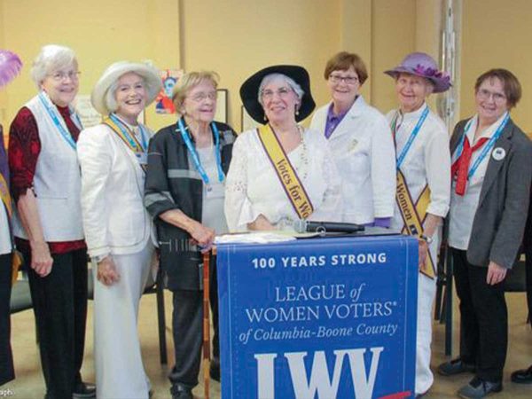 smiling. They are wearing various hats and sashes with the word Votes. They are posed behind a banner that reads 100 Years Strong