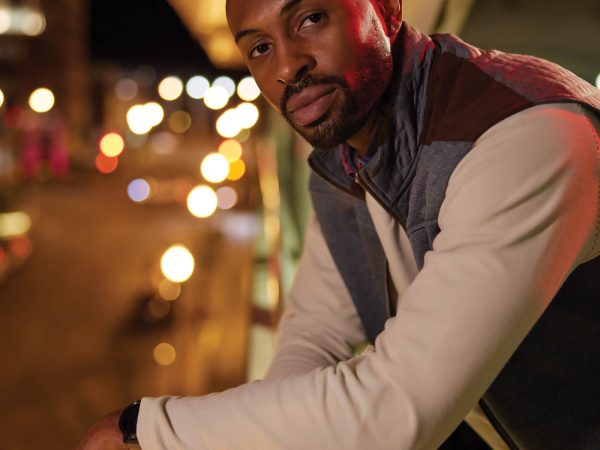 smiling at the camera. A sunset sky and city lights create a warm background.|Kari Hopkins|Kelly Hill|Marcus Jones|Megan Thomas|Megan Walters|Man in a suit with a serious expression is illuminated by red and blue lights against a blurred background of city lights