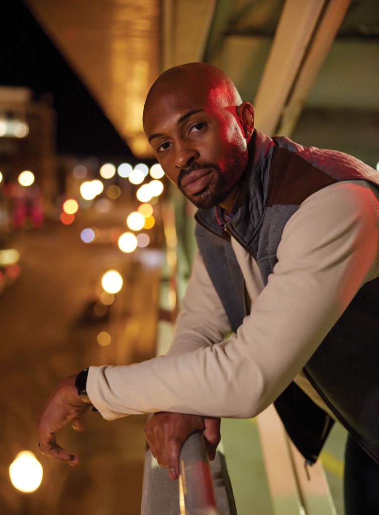 smiling at the camera. A sunset sky and city lights create a warm background.|Kari Hopkins|Kelly Hill|Marcus Jones|Megan Thomas|Megan Walters|Man in a suit with a serious expression is illuminated by red and blue lights against a blurred background of city lights