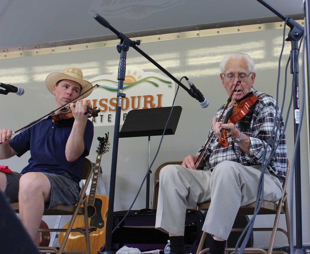 preforming and teaching her two apprentices inside of her home studio|three women sitting in wooden folding chairs in front of two microphones speaking on stage|an eldery musician playing a violin on stage next to a young man playing a violin|A female master artist in Colombian Dance