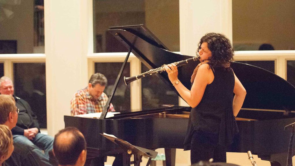 seated and attentive.|A pianist with long hair in a bun plays a grand piano on stage in a dimly lit venue. An audience sits at round tables in the foreground