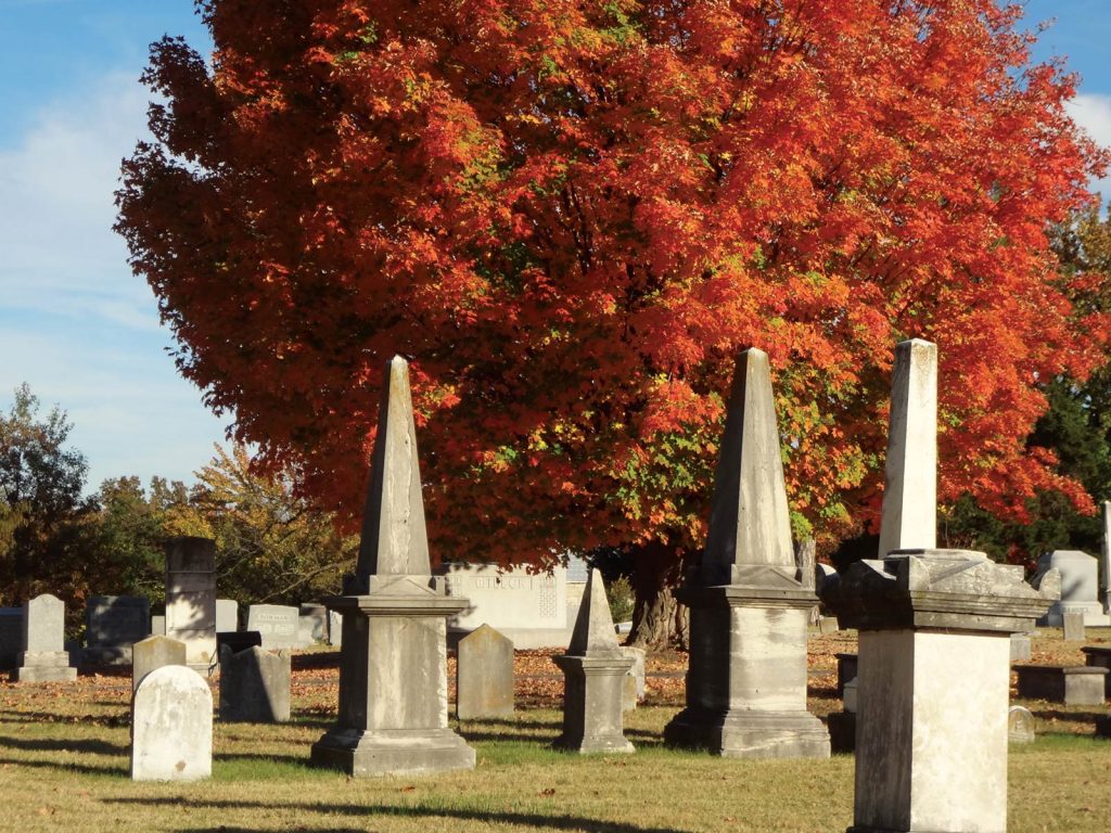 pointed tombstones set against a vibrant backdrop of a tree with bright orange and red autumn leaves. The sky is clear and blue