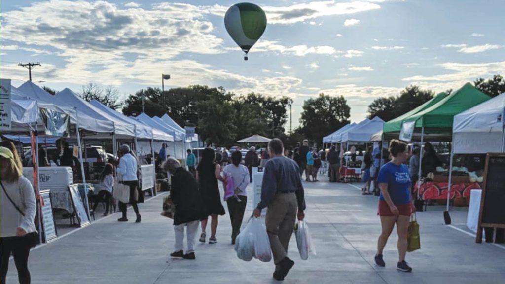 and others stroll under a partly cloudy sky. A hot air balloon floats in the distance. Trees border the scene.|A vendor in a black apron and cap stands at a market stall featuring various packaged food items and a blackboard menu. The background includes a van and a building under construction