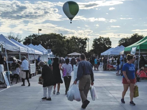 and others stroll under a partly cloudy sky. A hot air balloon floats in the distance. Trees border the scene.|A vendor in a black apron and cap stands at a market stall featuring various packaged food items and a blackboard menu. The background includes a van and a building under construction