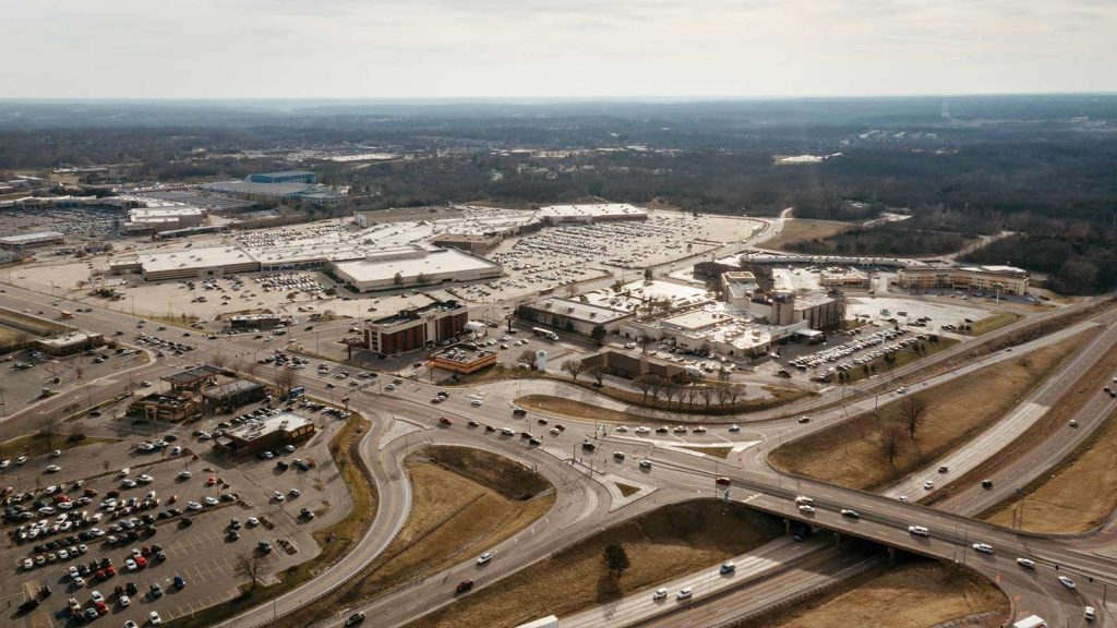 crisscrossed by busy highways and roads. Cars are visible on the roads and parked in the lot. The background shows a vast expanse of trees under a cloudy sky.|Illustration of a modern building with a mix of brick and glass facade. The building features multiple levels