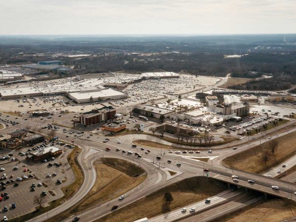 crisscrossed by busy highways and roads. Cars are visible on the roads and parked in the lot. The background shows a vast expanse of trees under a cloudy sky.|Illustration of a modern building with a mix of brick and glass facade. The building features multiple levels