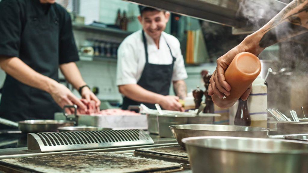 and various cooking utensils are visible.|Chefs in a commercial kitchen prepare ingredients. One holds a large piece of raw meat while another watches. Various cooking utensils and spices are on the counter. A third person stands nearby. The kitchen has a modern