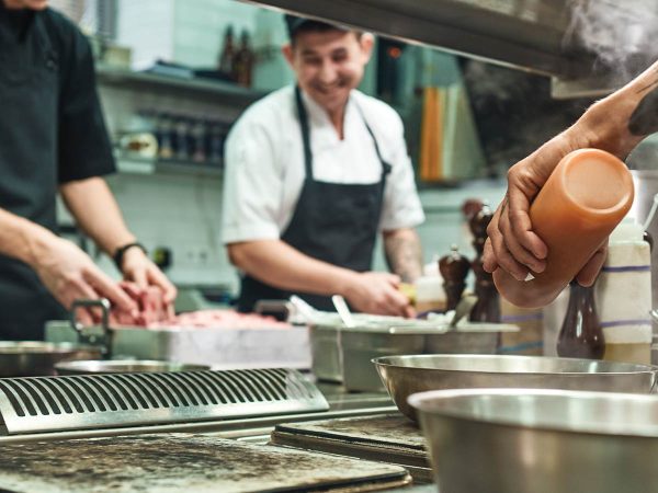 and various cooking utensils are visible.|Chefs in a commercial kitchen prepare ingredients. One holds a large piece of raw meat while another watches. Various cooking utensils and spices are on the counter. A third person stands nearby. The kitchen has a modern