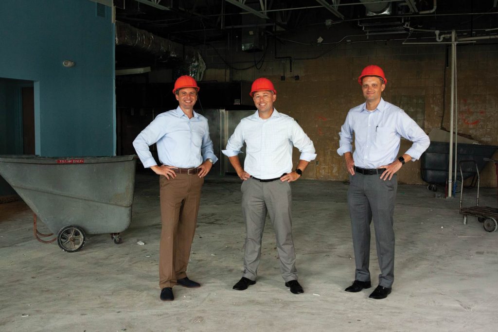 surrounded by construction materials and equipment.|A group of fourteen people stands outdoors in front of a brick building with arched windows. They are dressed in business casual attire